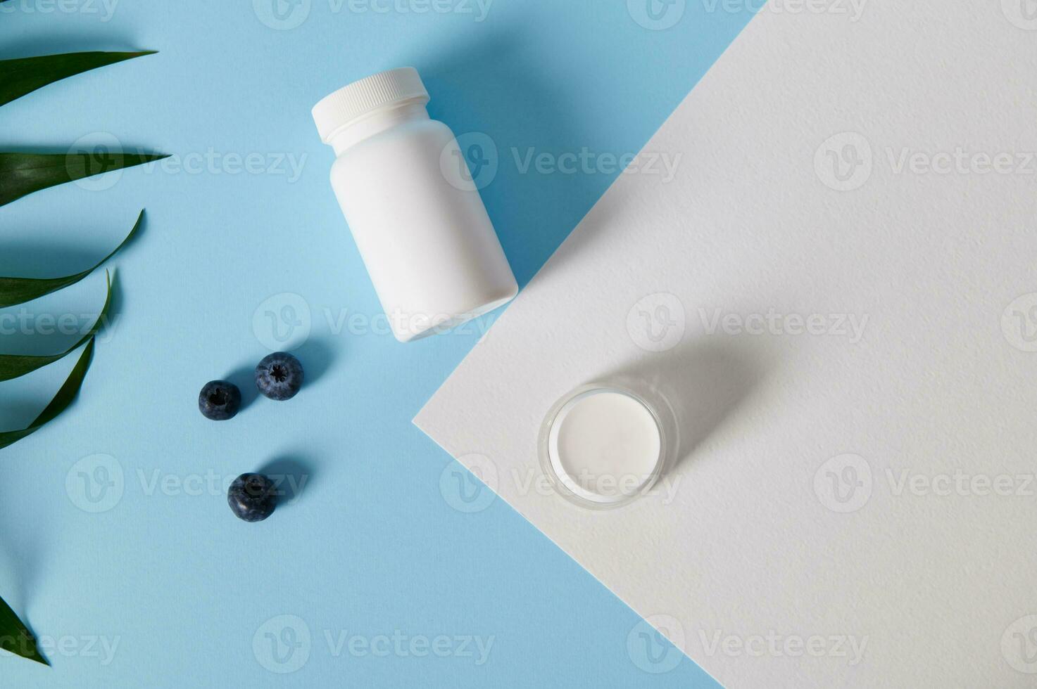 Flat lay of a pill container, bottle and scattered blueberries near a palm leaves on bicolor blue and white background photo