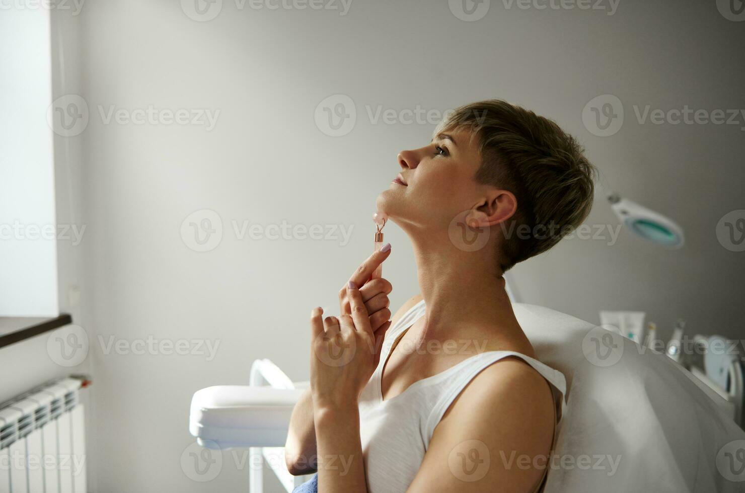 Side view of attractive blonde woman performing lymphatic drainage neck massage with jade stone roller massager photo