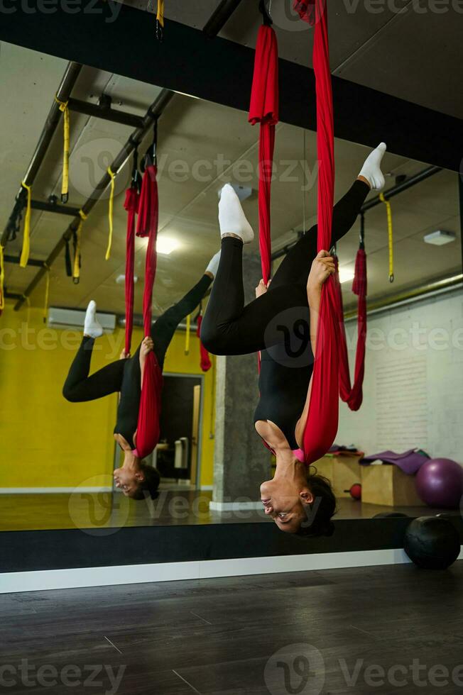Young African American fit woman practicing aerial flying yoga, doing stretching exercises hanging upside down on red hammock in yoga class photo