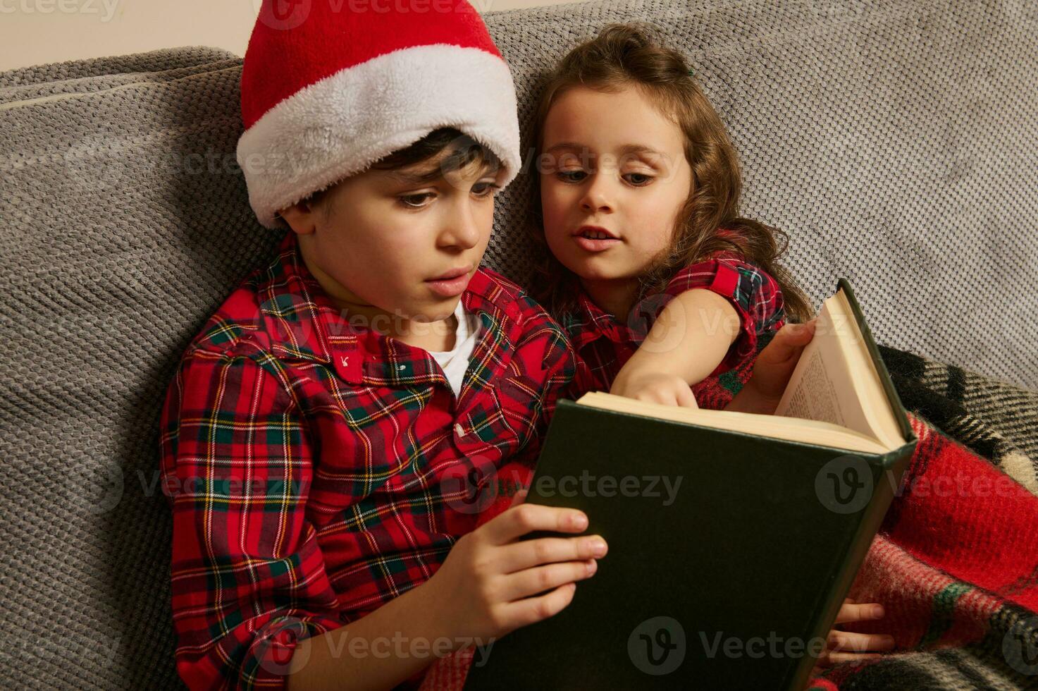 adorable europeo niños, hermano en de santa sombrero y su mas joven hermana sentado en el sofá y leyendo un libro, hada cuentos en el Navidad noche a hogar foto