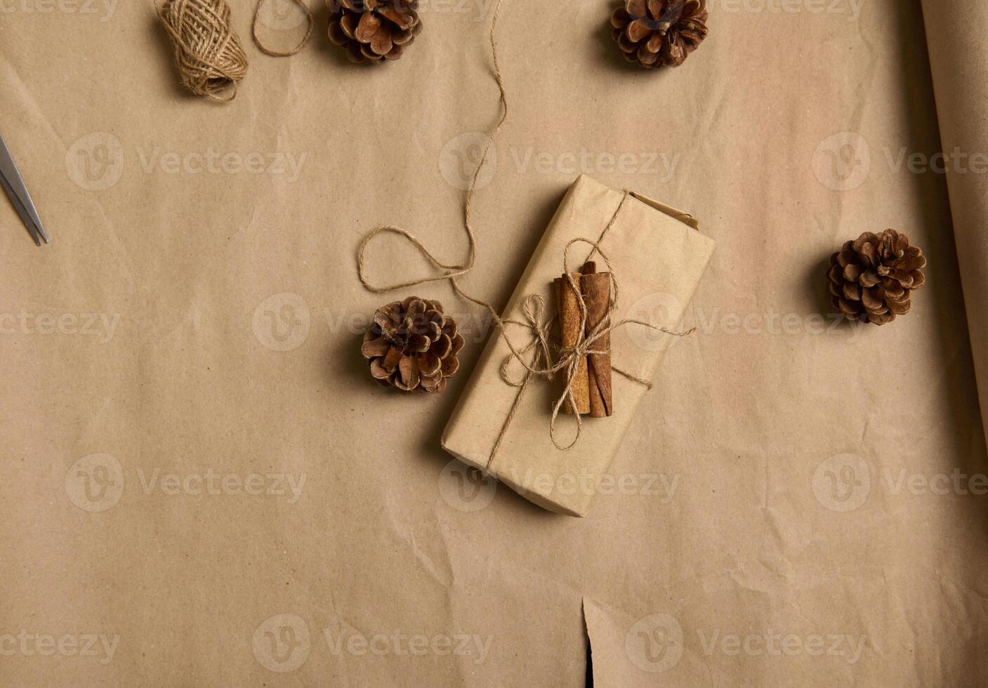 Flat lay minimalistic composition of a Christmas present wrapped in brown craft gift wrapping paper with a bow made of rope lying next to Christmas tree cones on a cardboard background with copy space photo