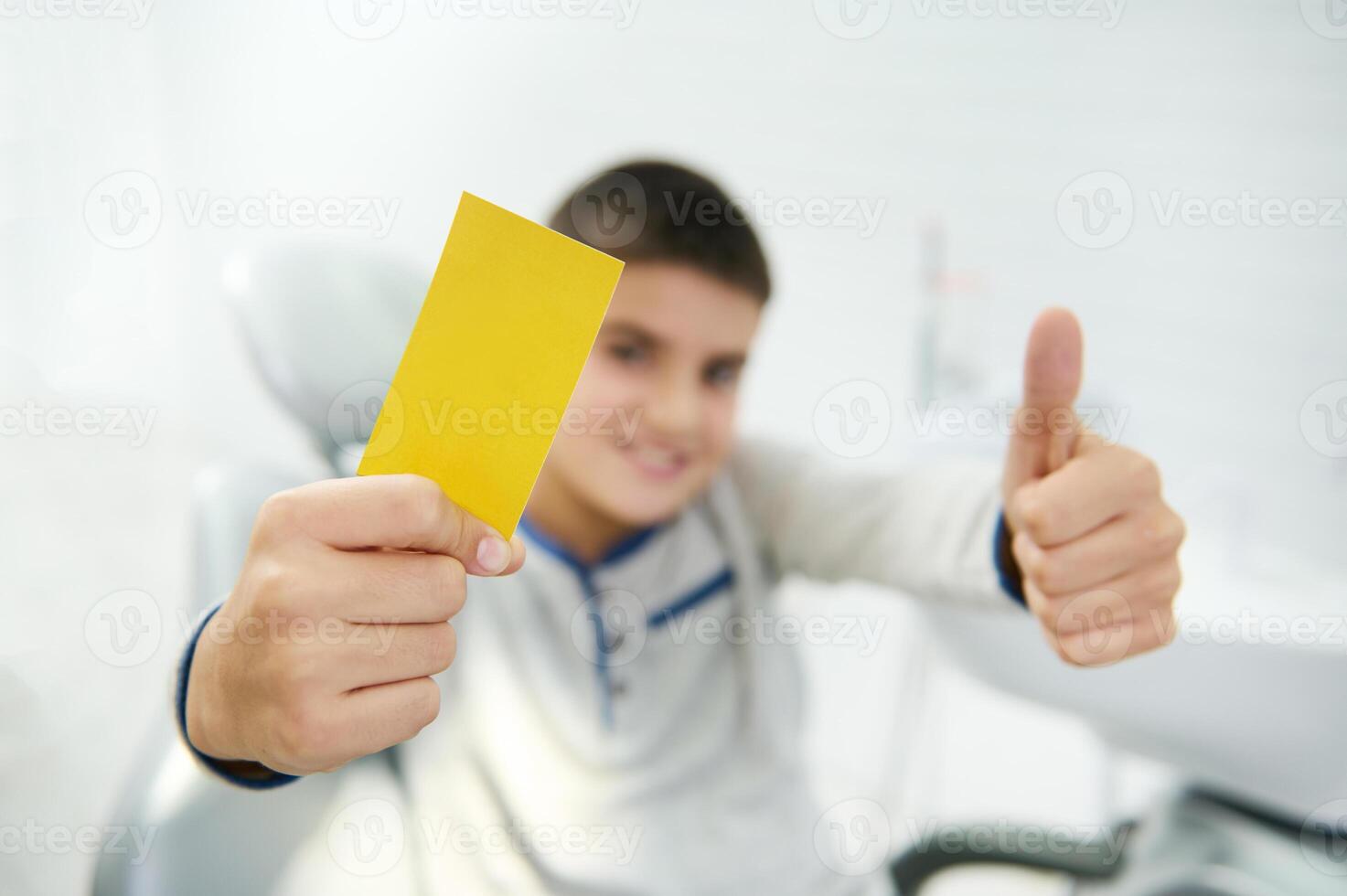 Focus on blank yellow visit card with copy space for insert ads and text in the hand of blurred handsome boy smiling toothy smile and showing thumb up looking at camera sitting in dentist chair photo