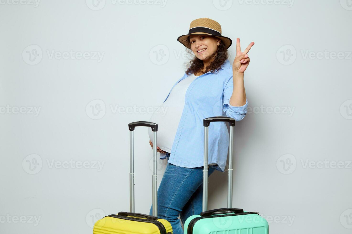 Happy pregnant woman in straw hat, caressing belly, showing peace sign, posing with colorful vibrant suitcases on white photo