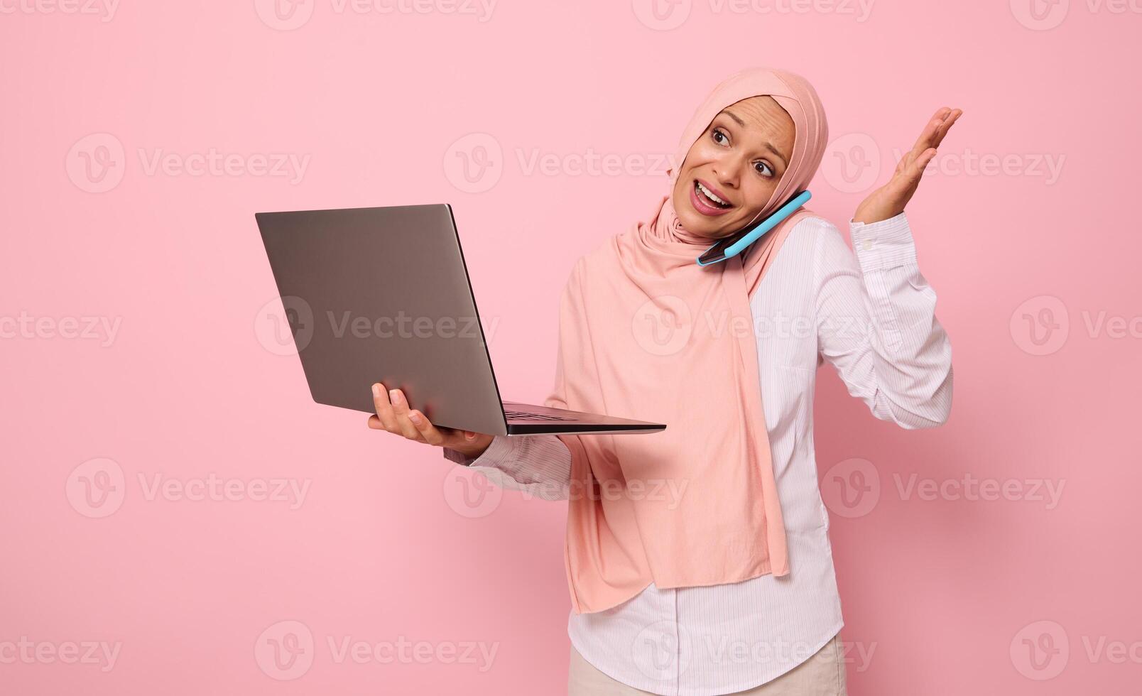 Busy and puzzled Muslim Arab pretty gorgeous woman in hijab, dressed business smart casual holds a laptop and talks on mobile phone , gestures with hands. Isolated over pink background with copy space photo