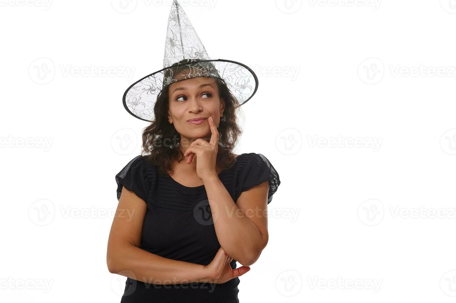 Charming Hispanic brunette in witch carnival hat and black outfit, holds finger on her chin and smiles with a cute toothy smile, staring mysteriously at copy space white background. Halloween concept photo