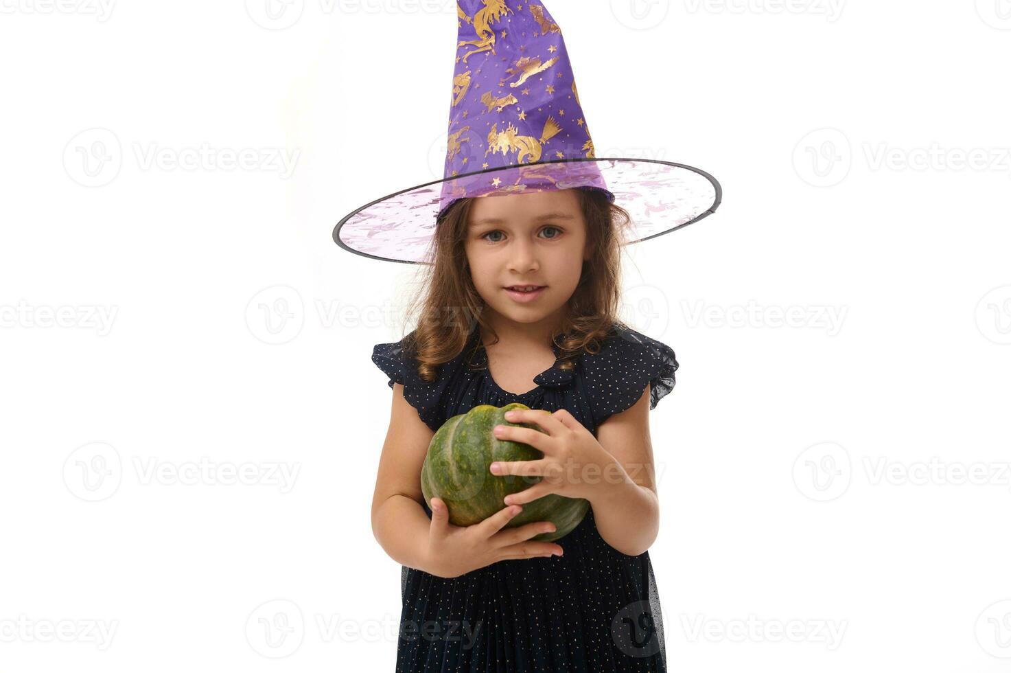 longitud de la cintura Disparo de pequeño caucásico niña vestido en elegante carnaval atuendo y mago sombrero, bruja niño posando con calabaza en contra un blanco antecedentes con Copiar espacio. Víspera de Todos los Santos bruja concepto foto