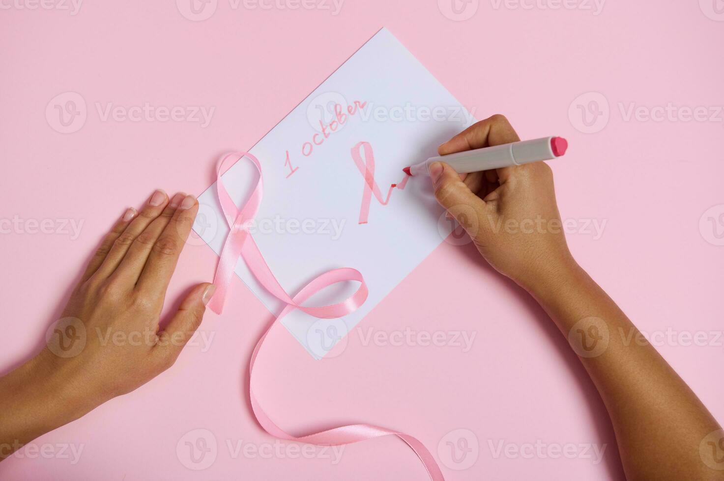 Top view of a woman's hand holding a felt-tip pen writes october 1 and draws on the paper a pink symbol of breast cancer awareness month, a pink ribbon with an endless end, lying on a pink background photo