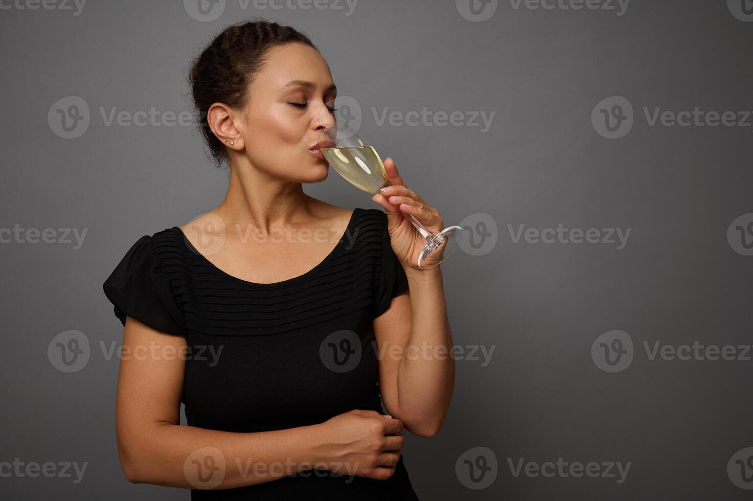 Cheerful woman in black outfit drinks sparkling wine from a glass, standing isolated over gray wall background with copy ad space photo