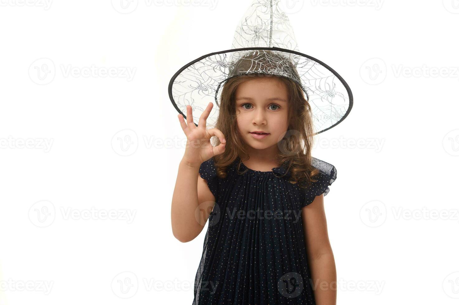 Portrait of pretty little witch girl wearing a wizard hat and dressed in stylish carnival dress, gesturing, showing OK sign with fingers. Halloween concept on white background with copy space photo