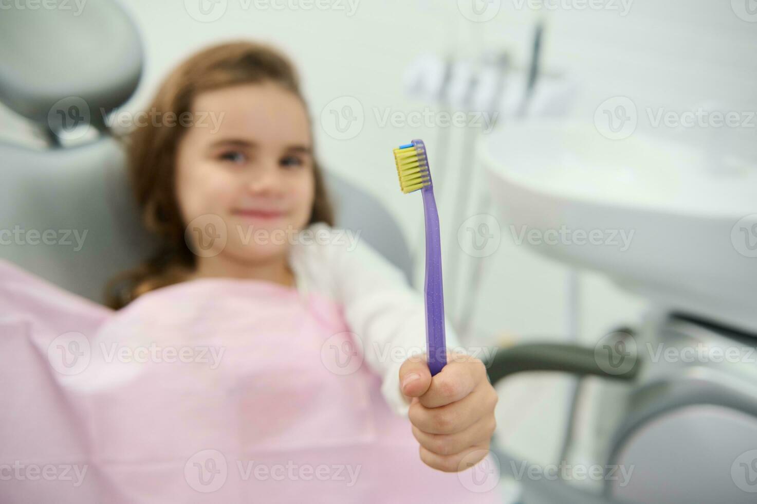 atención en un cepillo de dientes en el mano de un alegre bonito niña lindo sonriente mirando a cámara sentado en del dentista silla durante dental chequeo. oral cuidado y higiene. pediatra odontología concepto foto