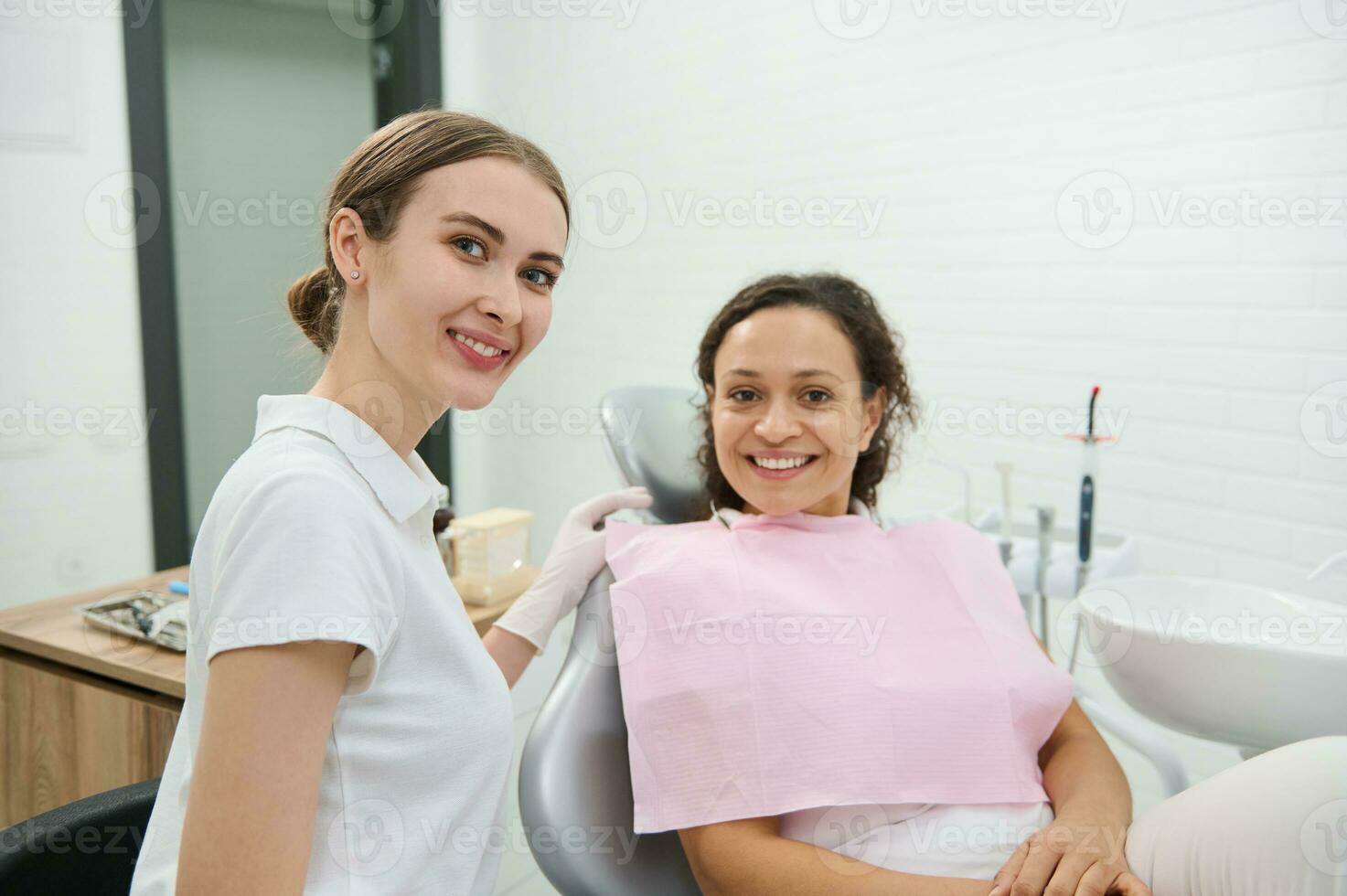 Attractive confident woman, dentist hygienist cutely smiles looking at camera beside female patient sitting in dentists chair before receiving dental treatment in dentistry clinic. Oral hygiene care photo