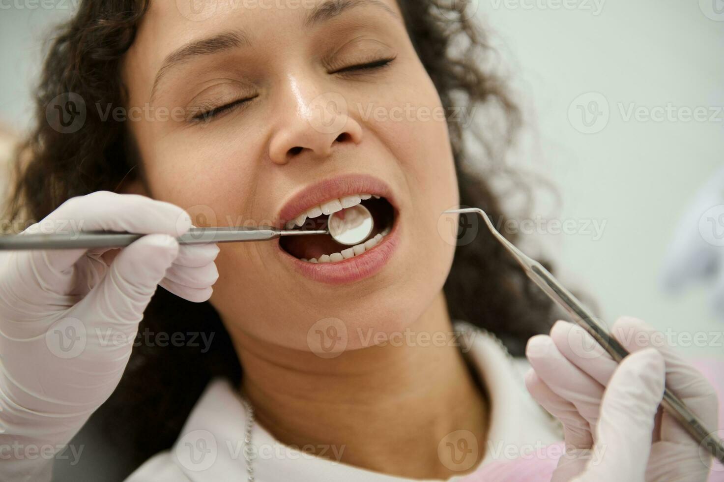 de cerca de bonito mujer, paciente en odontología clínica, con cerrado ojos y abierto boca mientras higienista dentista examinando su dientes con estéril inoxidable acero dental espejo. oral higiene concepto foto