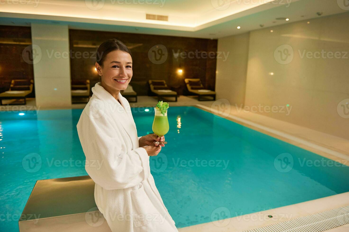 encantador mujer en blanco bata de baño soportes por el piscina en lujoso un bienestar spa complejo, sostiene recién exprimido vitamina jugo en manos, sonrisas mirando a cámara. sano estilo de vida, recreación concepto foto