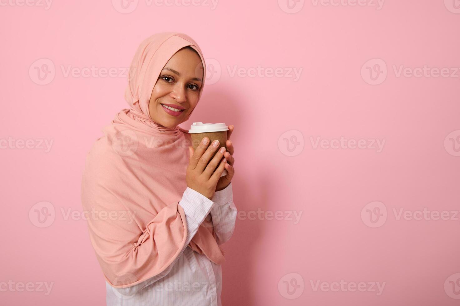 Arabic muslim pretty woman with covered head in pink hijab drinking hot drink, tea or coffee from disposable cardboard takeaway cup, standing three quarters against colored background with copy space photo