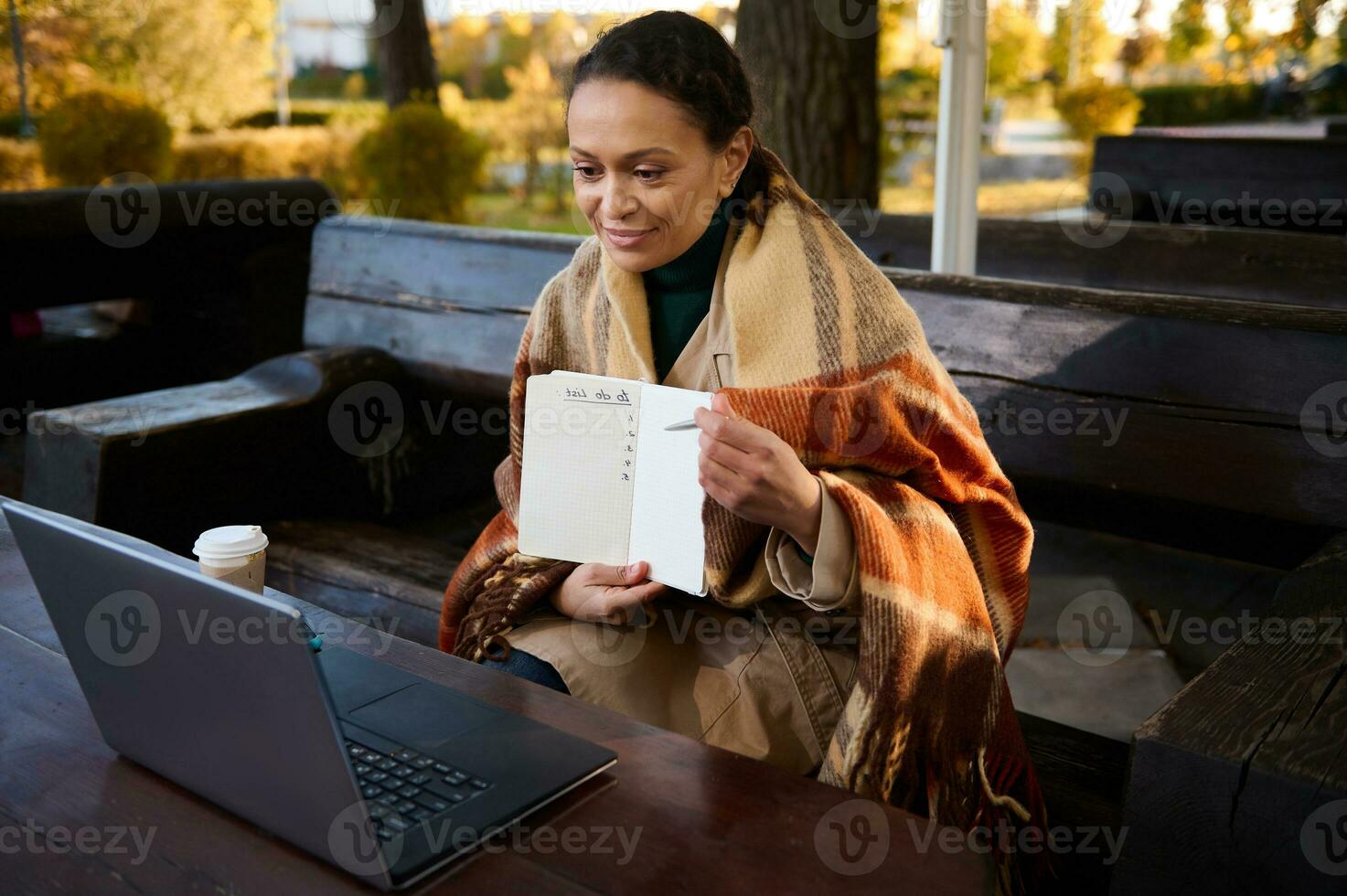 Pretty woman showing a blank page of copybook during video call on laptop, keeps warm by wrapping herself in woolen blanket, sitting outdoor on wooden bench. Diastant work, internet learning concept photo