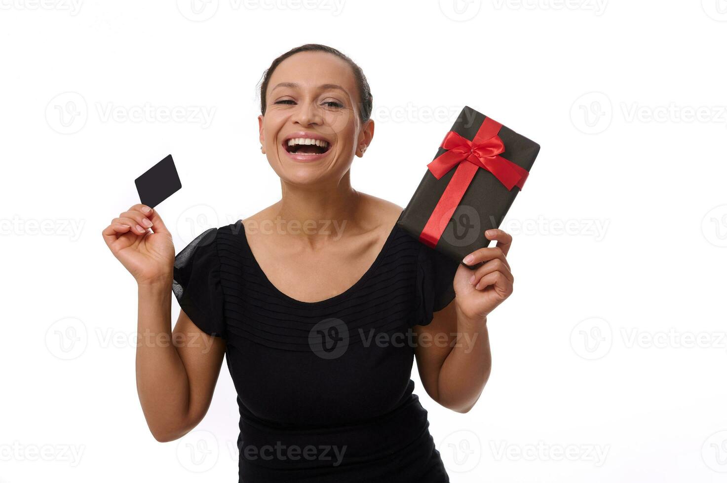 Isolated portrait on white background with copy space of attractive laughing African American woman dressed in black, holding discount credit card and gift box, looking at camera. Black Friday concept photo