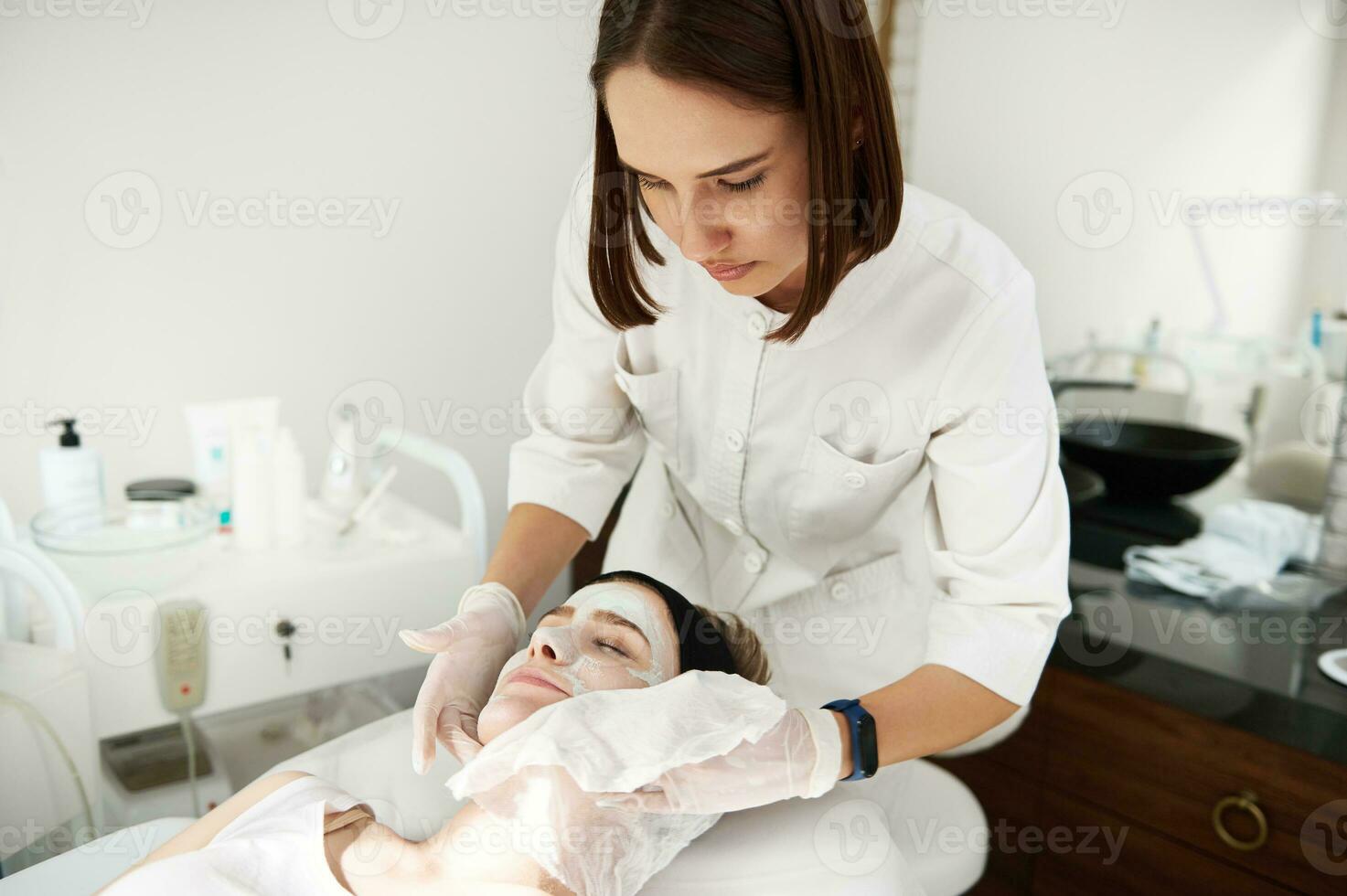 médico cosmetólogo utilizando servilletas para quitando belleza hidratar máscara desde mujer cara en spa salón. foto