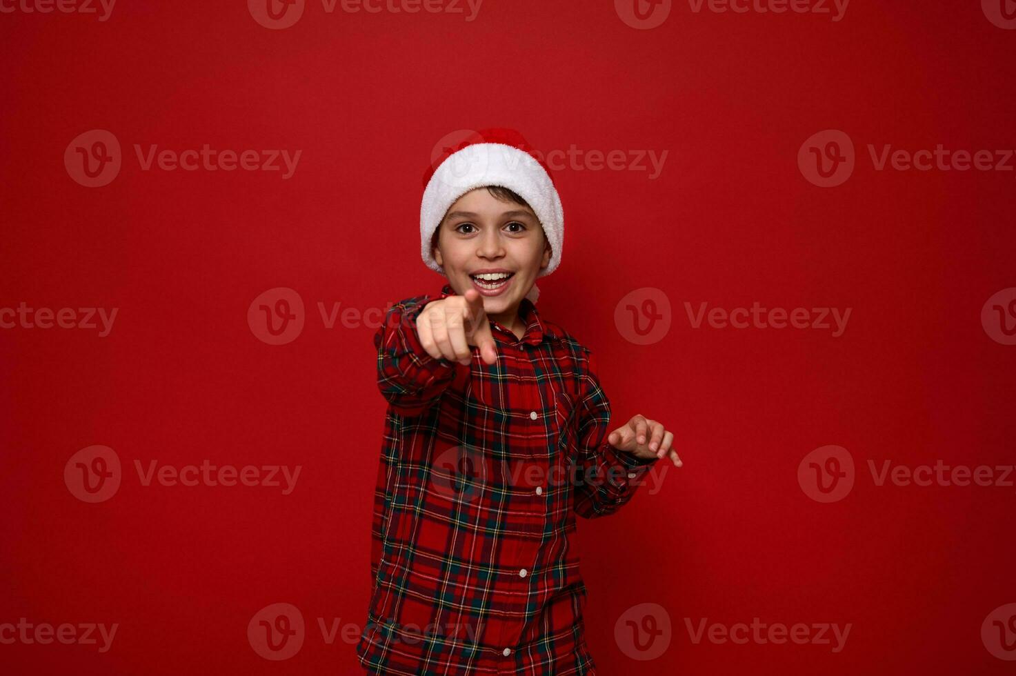 Adorable preadolescent boy in red checkered shirt wearing a Santa hat points directly to camera, smiles with beautiful toothy smile posing against colored background with copy space for Christmas ad photo