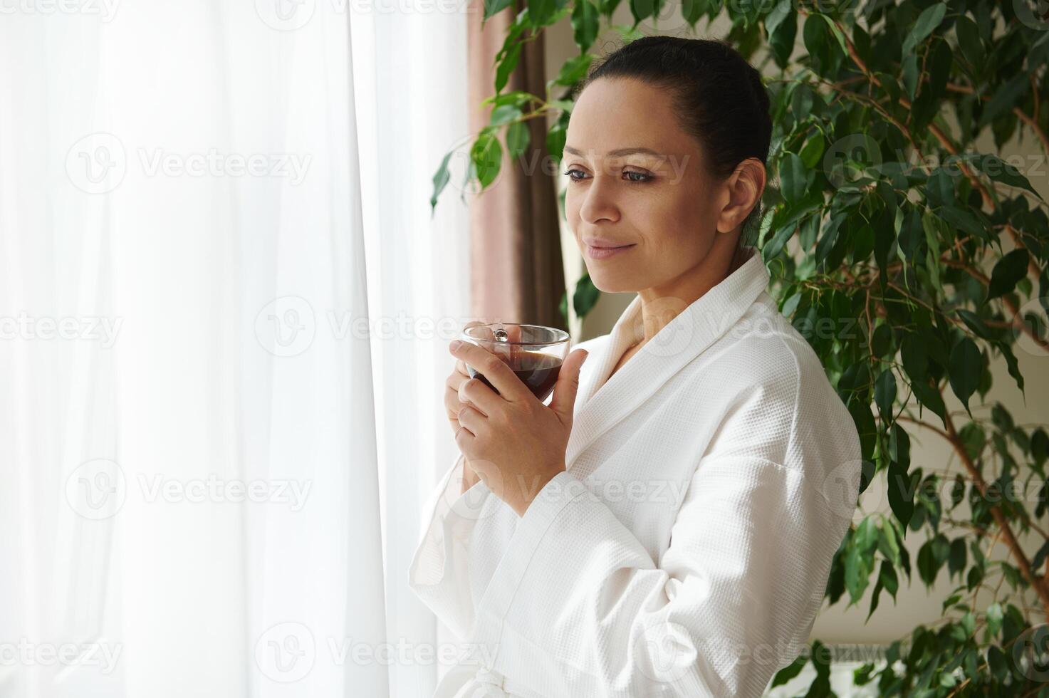 A serene mature woman in a white robe stands with a cup of coffee in her hands against the background of a houseplant and enjoys the view outside the window through transparent white curtains photo