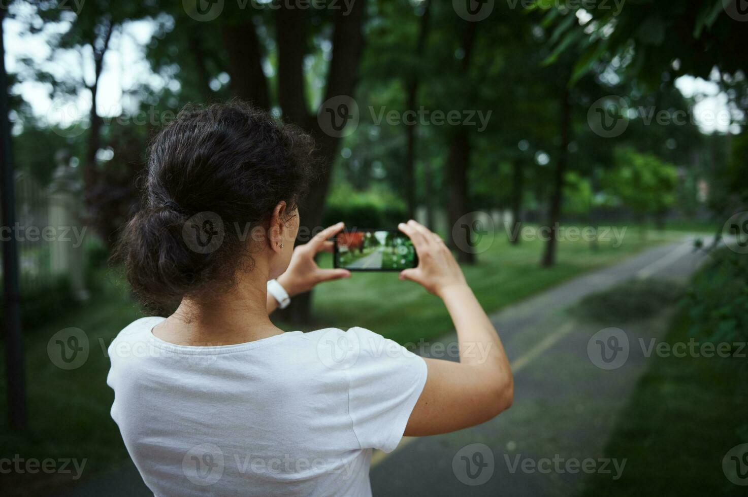 Rear view of a young woman holding mobile phone in live view mode camera, taking photo of the park alley