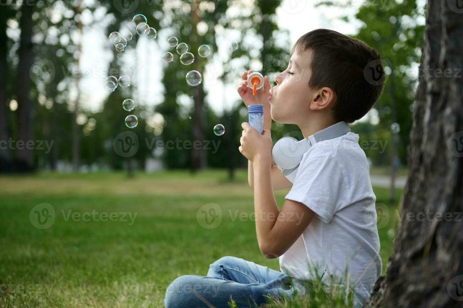 Close-up side portrait of an adorable cheerful beautiful cute elementary aged school boy enjoying his recreation between classes, blowing soap bubbles, sitting on the green grass of urban city park photo