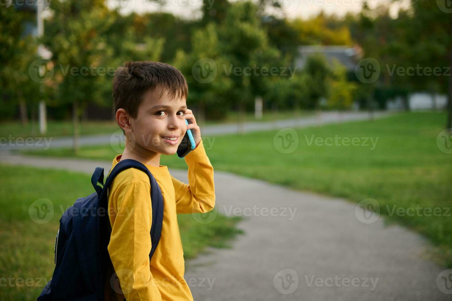 Side view of a schoolboy wearing yellow sweatshirt with backpack walking on the path in public park, going home after school, talking on mobile phone, smiling with toothy smile to the camera photo