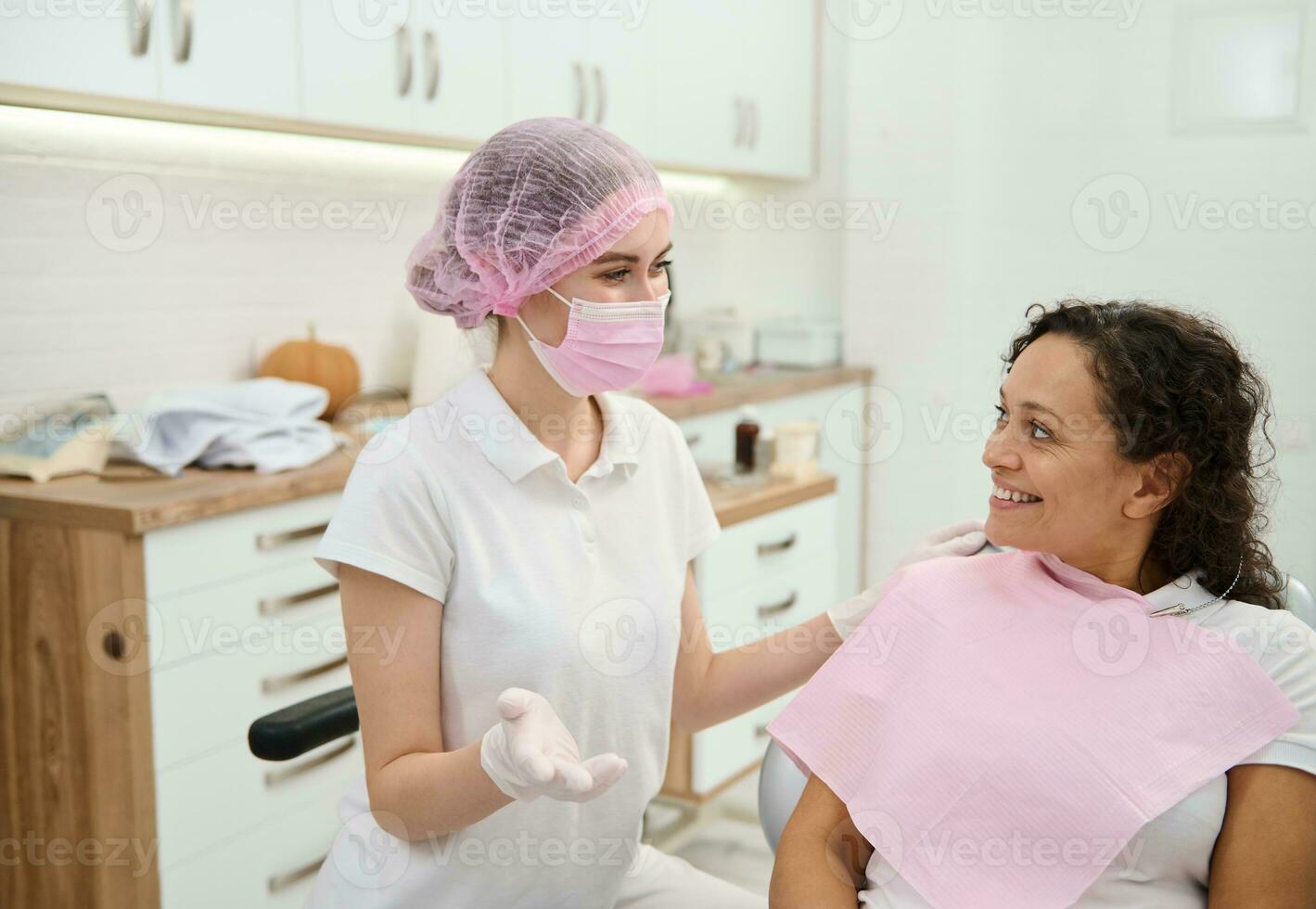 dentista higienista, dental asistente vistiendo rosado médico máscara y quirúrgico guantes preparando mujer paciente a médico chequeo, dando consulta acerca de oral higiene en odontología moderno clínica foto