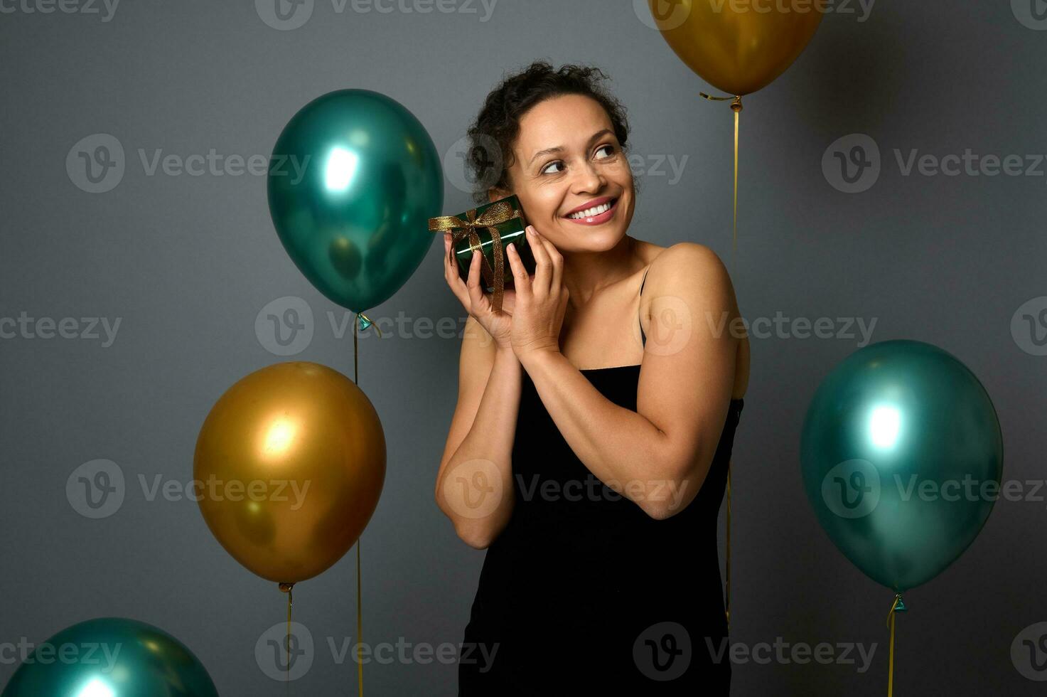 Joyful mixed race pretty woman with beautiful smile holds a gift box near her face, poses on gray background with golden green air balloons. Anniversary, Christmas, New Year concepts for advertising. photo