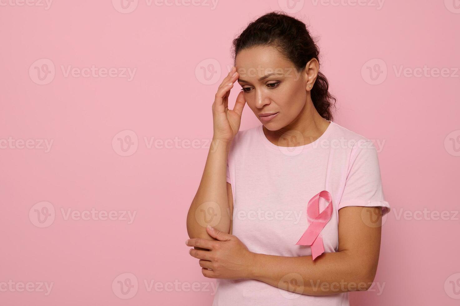 Beautiful thoughtful woman in pink t-shirt with pink ribbon looking down on pink background, hands on temple. International World Breast Cancer Awareness Day, Educational, medical concept, copy space photo