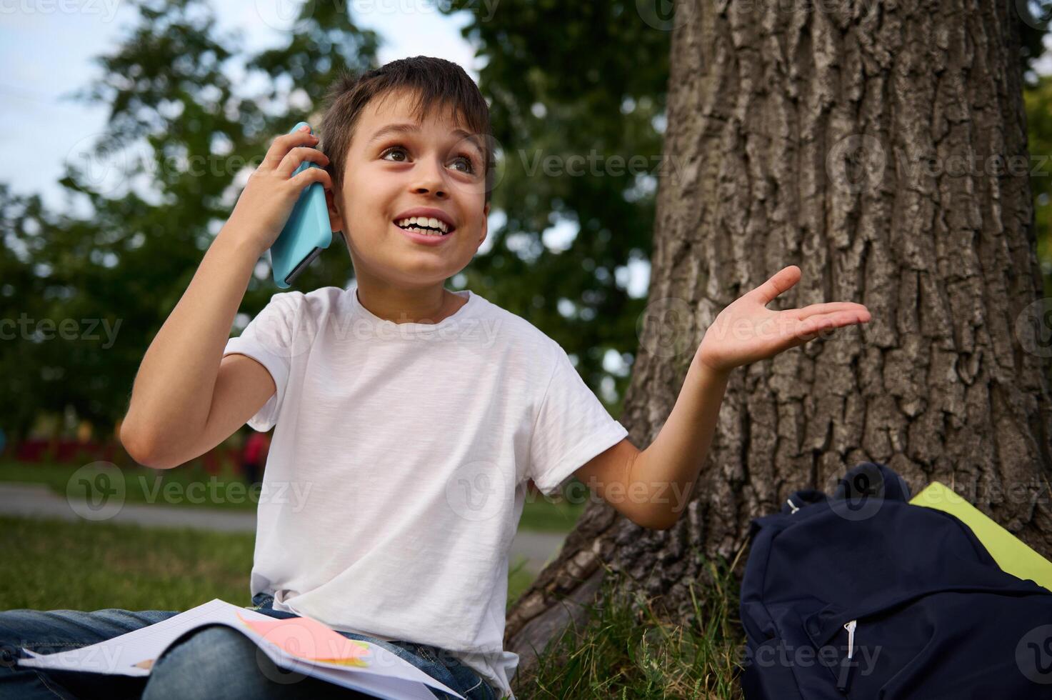 Cheerful adorable school child boy talking on mobile phone, cute smiling looking away, gesturing with hands, resting on the public park after first day at school on beautiful summer day. photo