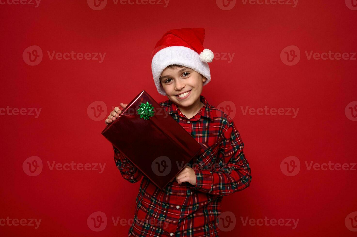 Handsome preadolescent boy in Santa Claus hat and red plaid shirt poses over colored background with Christmas gift, smiled with beautiful toothy smile looking at camera. Copy space for advertisement photo