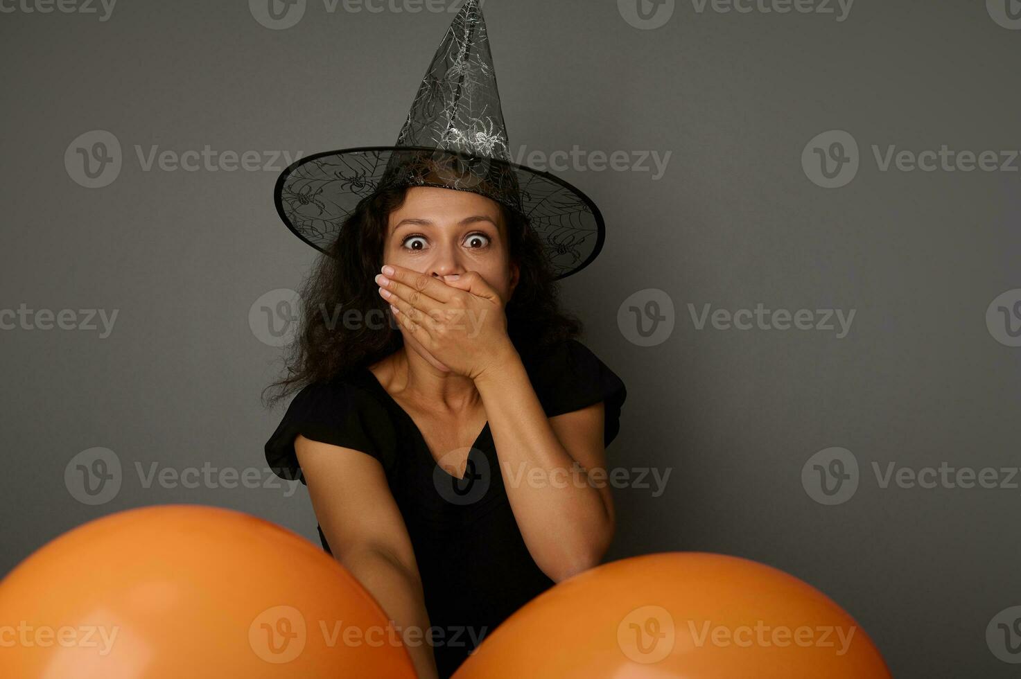 estupefacto morena mujer vestido en negro bruja carnaval atuendo cubre su boca desde horror, mira a cámara posando en contra un gris antecedentes con vistoso naranja aire pelotas. Víspera de Todos los Santos concepto foto