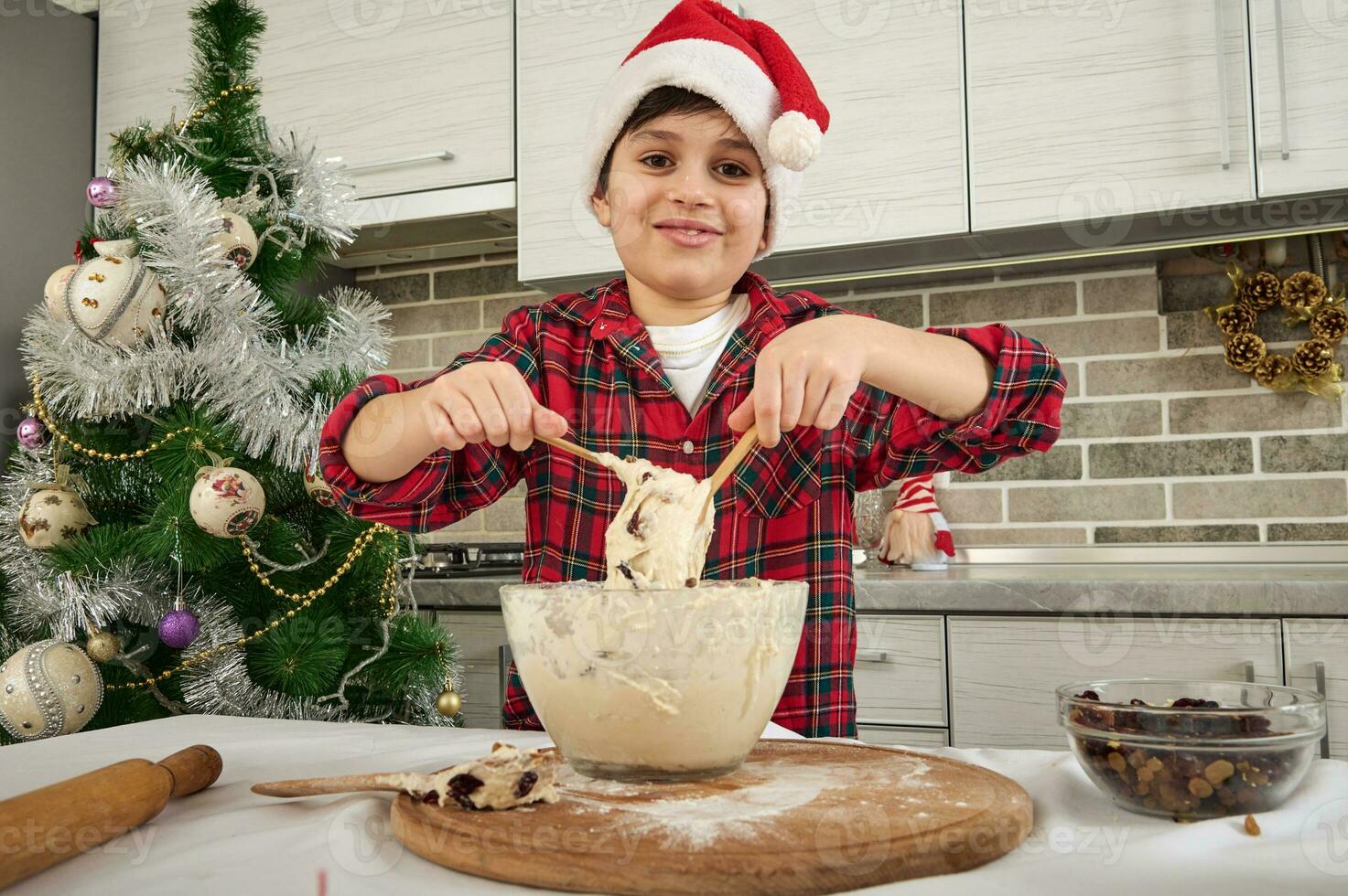 Handsome preadolescent Caucasian boy, adorable kid chef blogger in Santa hat kneading dough and showing it to the camera, cutely smiling with cheerful toothy smile. Christmas culinary concept photo