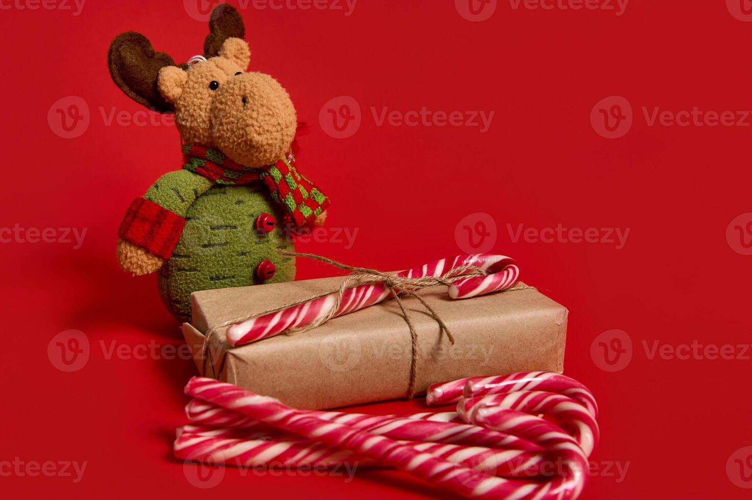 Beautiful minimalist composition of Christmas items, gift box in kraft wrapping paper with tied bow, sweet lollipops and deer plush toy on red background with copy space for ad photo