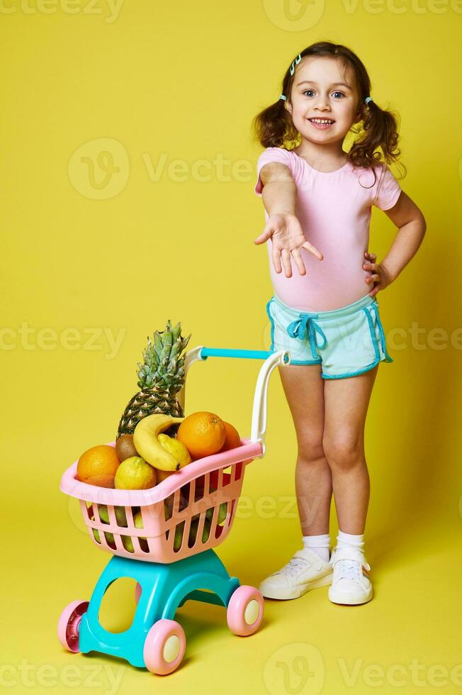 Beautiful little girl in summer clothes pointing her hand, showing a shopping cart full of delicious fruits. Isolated on yellow background, copy space photo