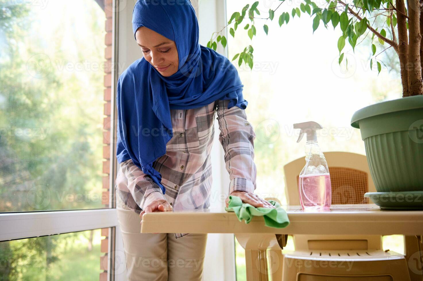 árabe musulmán mujer con cabeza cubierto en hijab haciendo limpieza el casa, limpiando apagado el polvo con un verde trapo desde un sucio mesa superficie en el veranda. limpieza interna, quehaceres, limpieza concepto foto