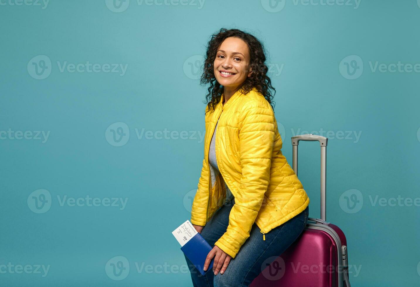 Beautiful woman in bright colorful clothes, with passport, air ticket and boarding pass, sitting on her suitcase and smiling toothy smile looking at camera. Travel. Tourism concept. Blue background photo