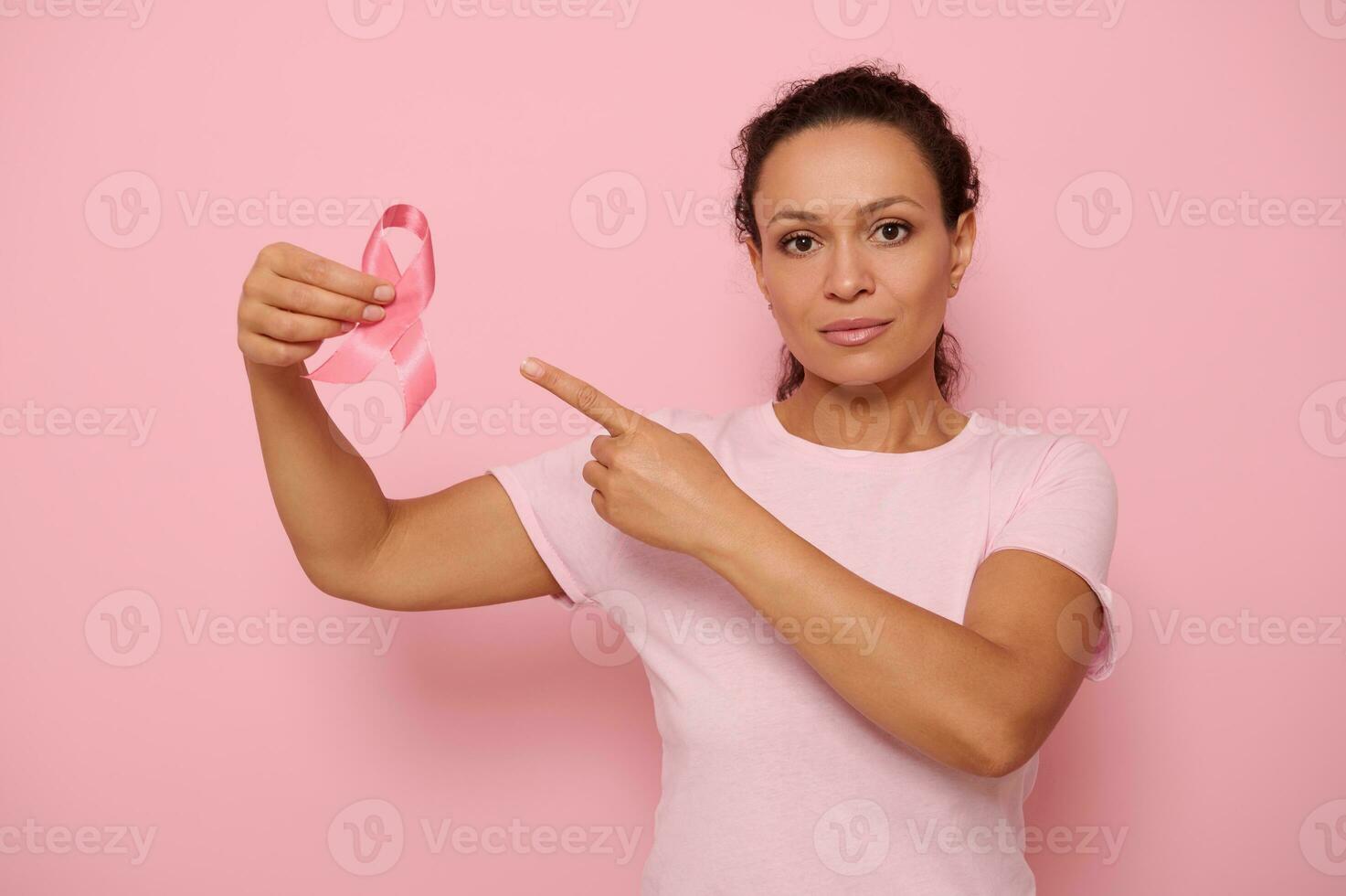 Portrait of serene Middle Eastern ethnicity woman in pink T-shirt pointing to a satin ribbon in her hand, looking at camera, isolated on colored background with copy space. Breast Cancer Day concept photo