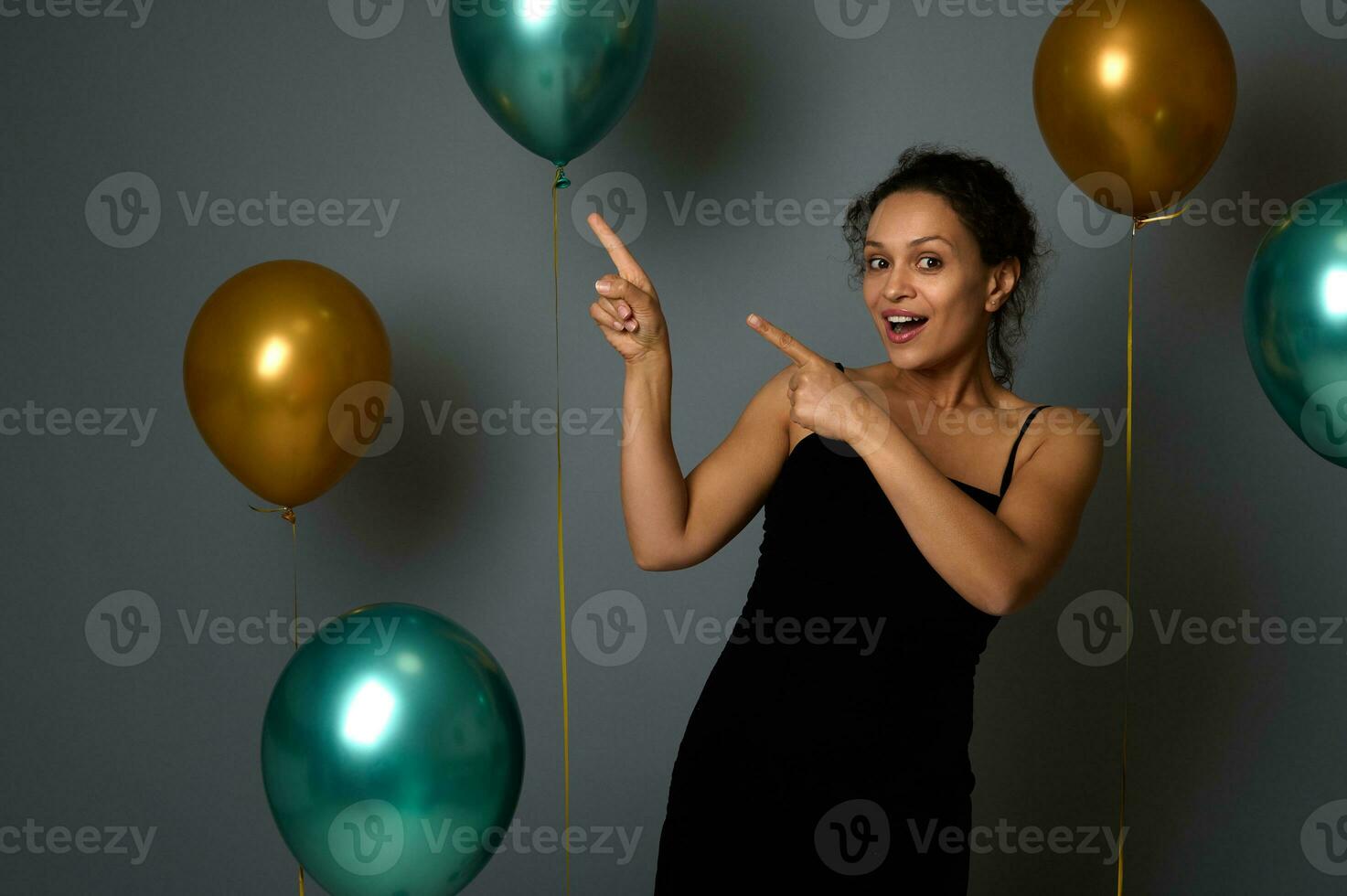 Attractive joyful woman in evening attire looks at camera and points on a gray background copy space decorated with golden green air balloons for Christmas, anniversary , New Year advertisement photo