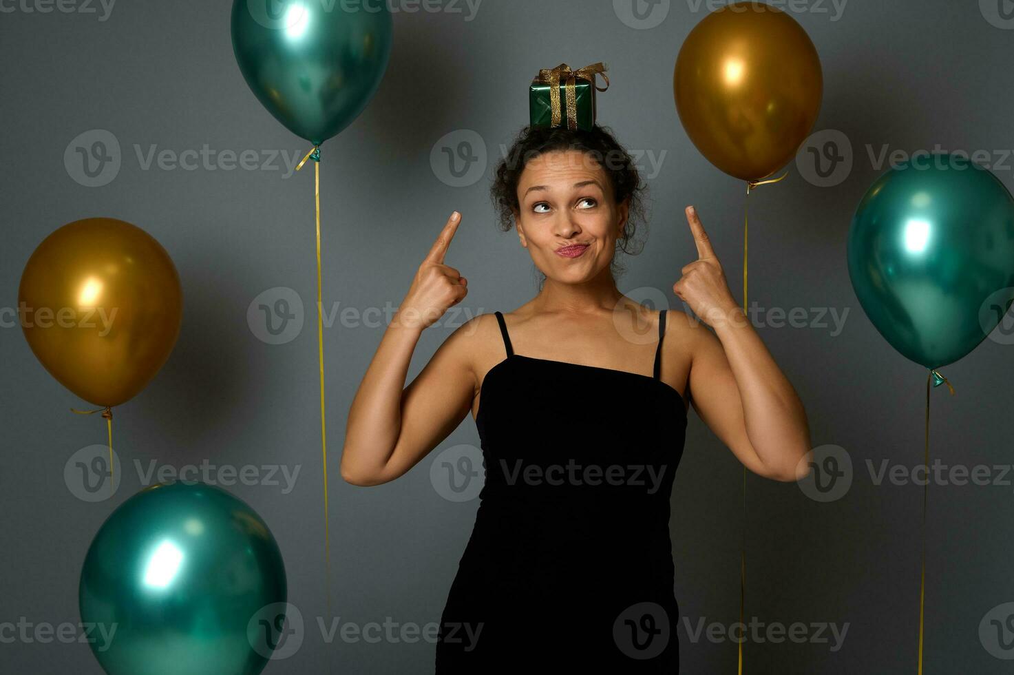 African pretty woman in evening dress points fingers on a Christmas gift on her head, looks up, poses against gray wall background decoration with golden green metallic air balls. Celebration concept photo