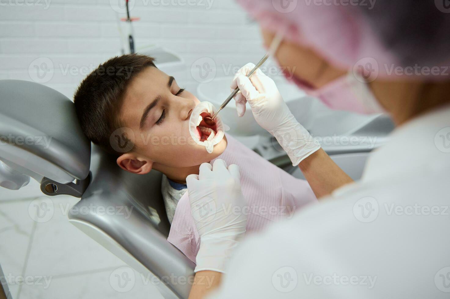 Confident child boy sitting in dentist's chair in contemporary dental clinic while unrecognizable female dentist doing medical manipulations in his opened mouth by cheek retractor and dental mirror photo