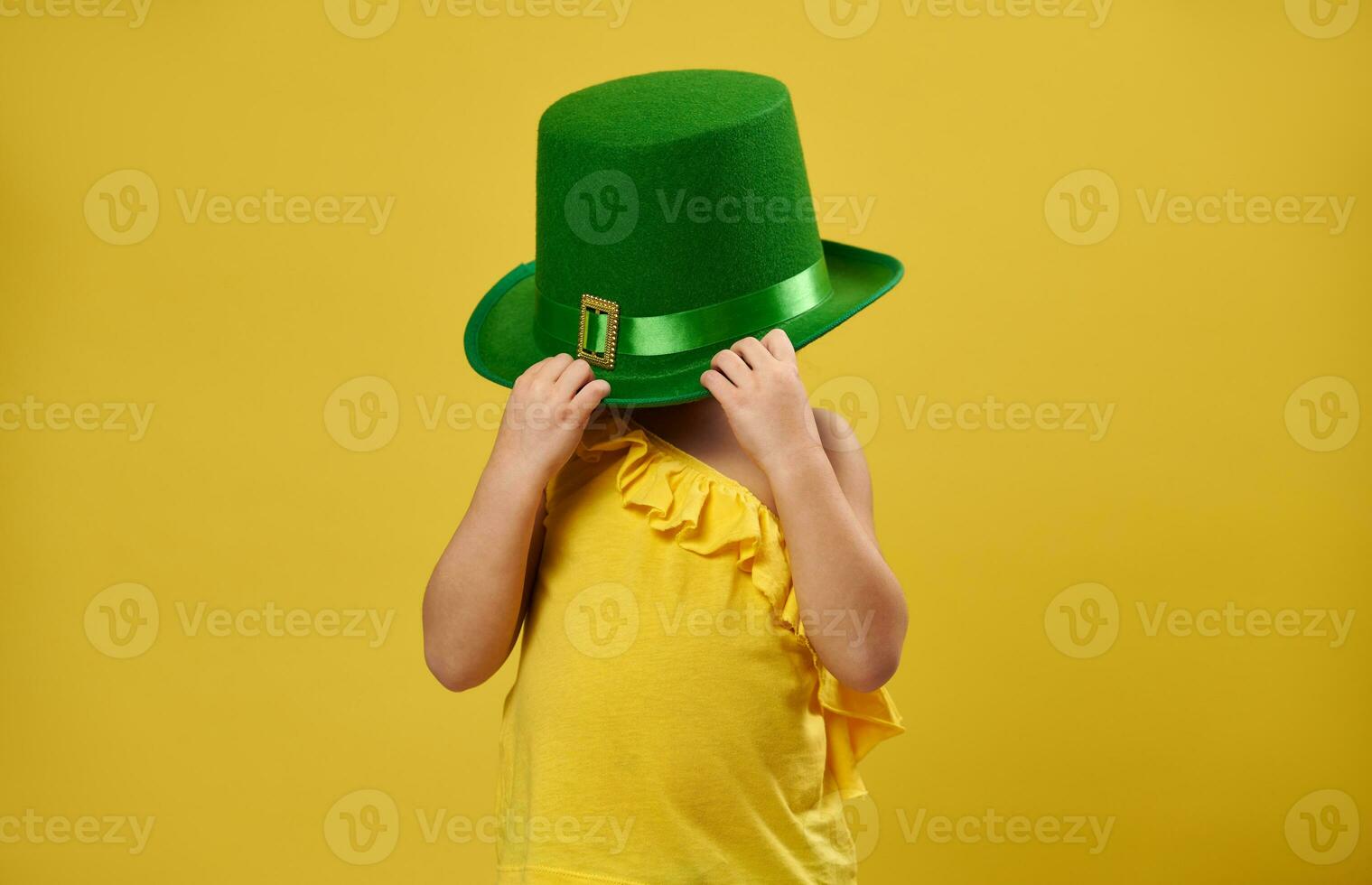 Little girl hides her face under a green Irish leprechaun hat. Saint Patrick's Day concept on yellow background. photo