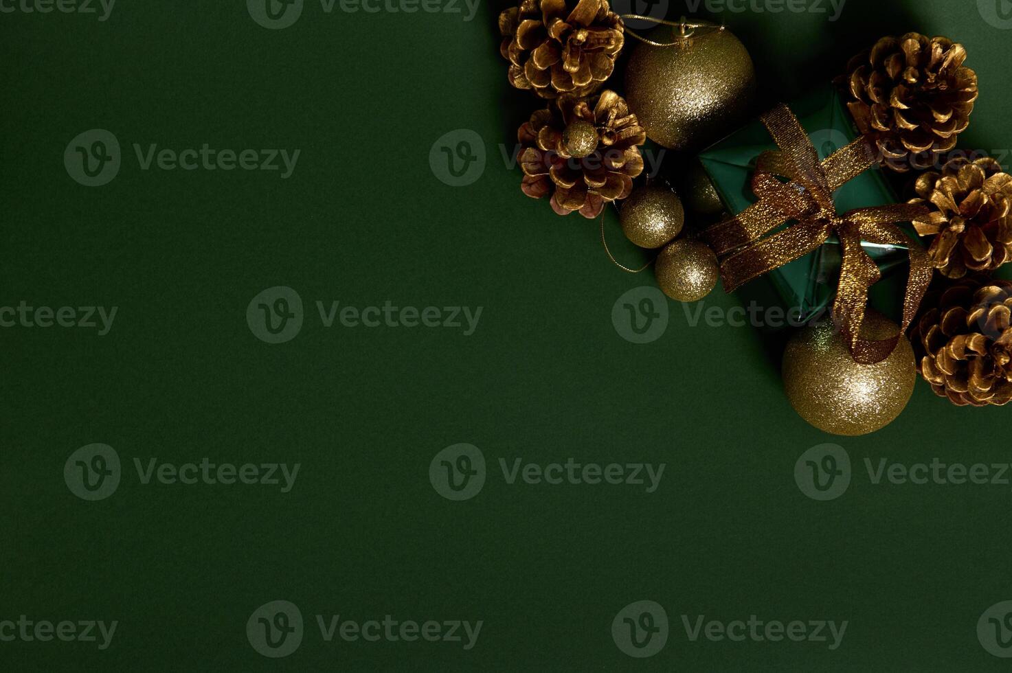 Flat lay of a present in shiny wrapping gift paper with golden bow, pines cones and gold Christmas tree toys laid out on the corner of green background. Copy space for advertisement photo