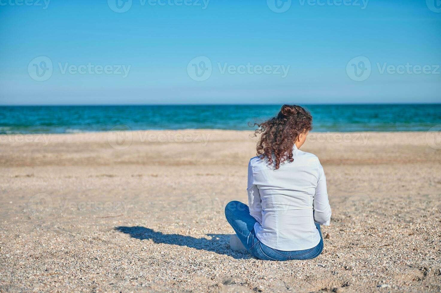 Rear view of a young woman in white shirt and blue jeans sitting on the sand in the beach on a beautiful sunny day photo