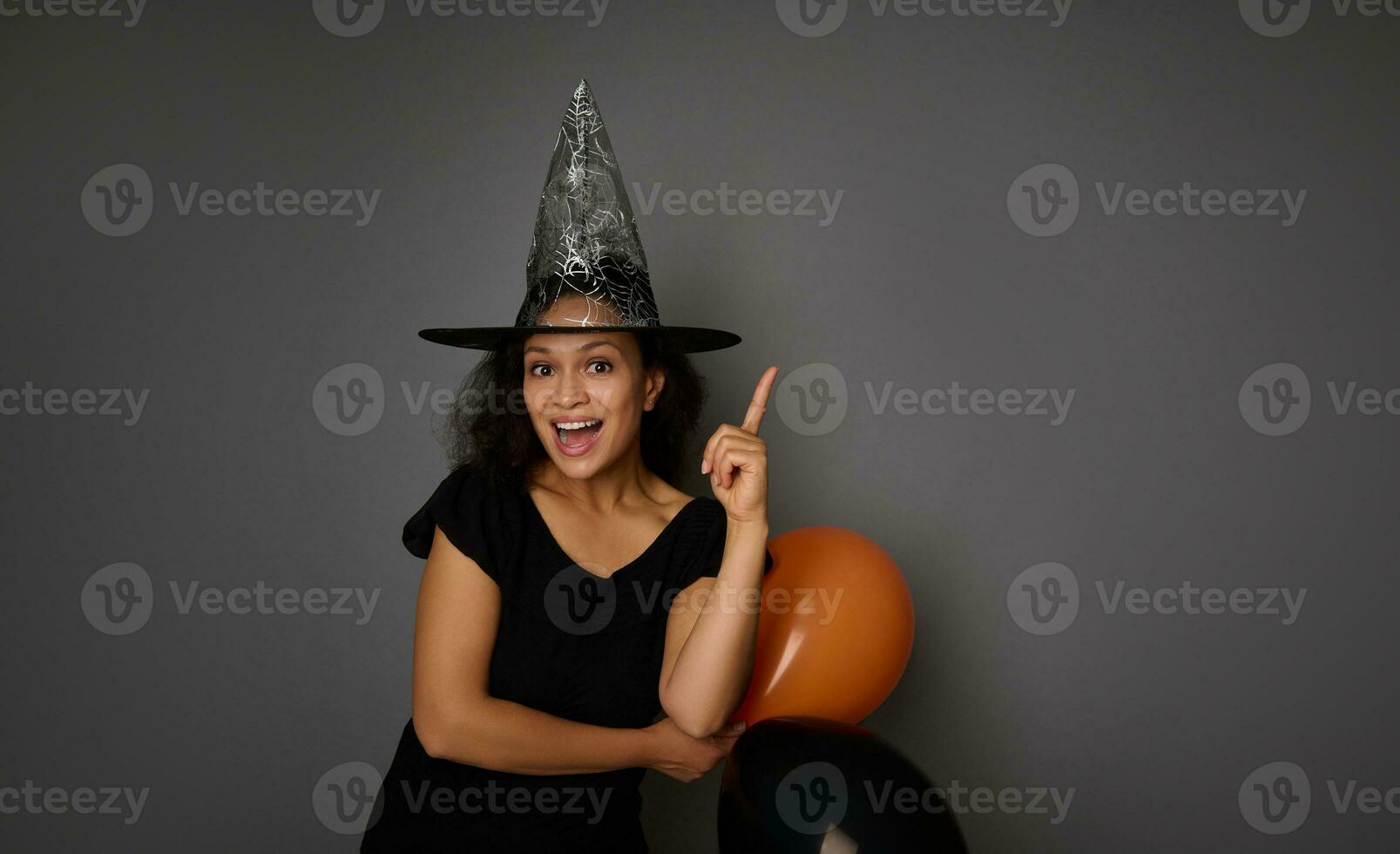 Cheerful Hispanic woman in wizard hat, dressed in black, holds two orange and black air balloons and points on a copy space on gray background. Halloween party concept photo