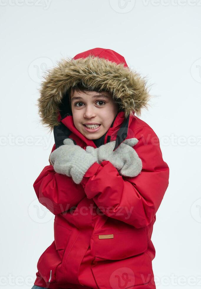 Isolated portrait on a white background with copy ad space of a handsome European boy, dressed in a bright red down jacket with hood, looking at the camera while hugging himself photo