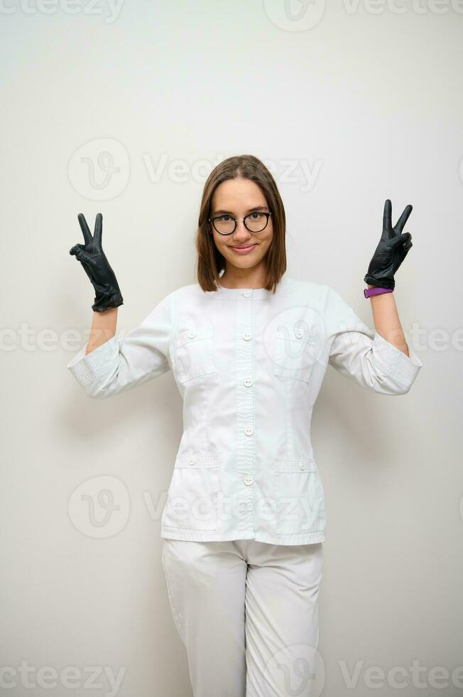 hermosa joven morena mujer, médico interno, hembra médico en blanco uniforme sonrisas mirando a cámara y demostración paz firmar en contra blanco pared antecedentes con Copiar espacio foto