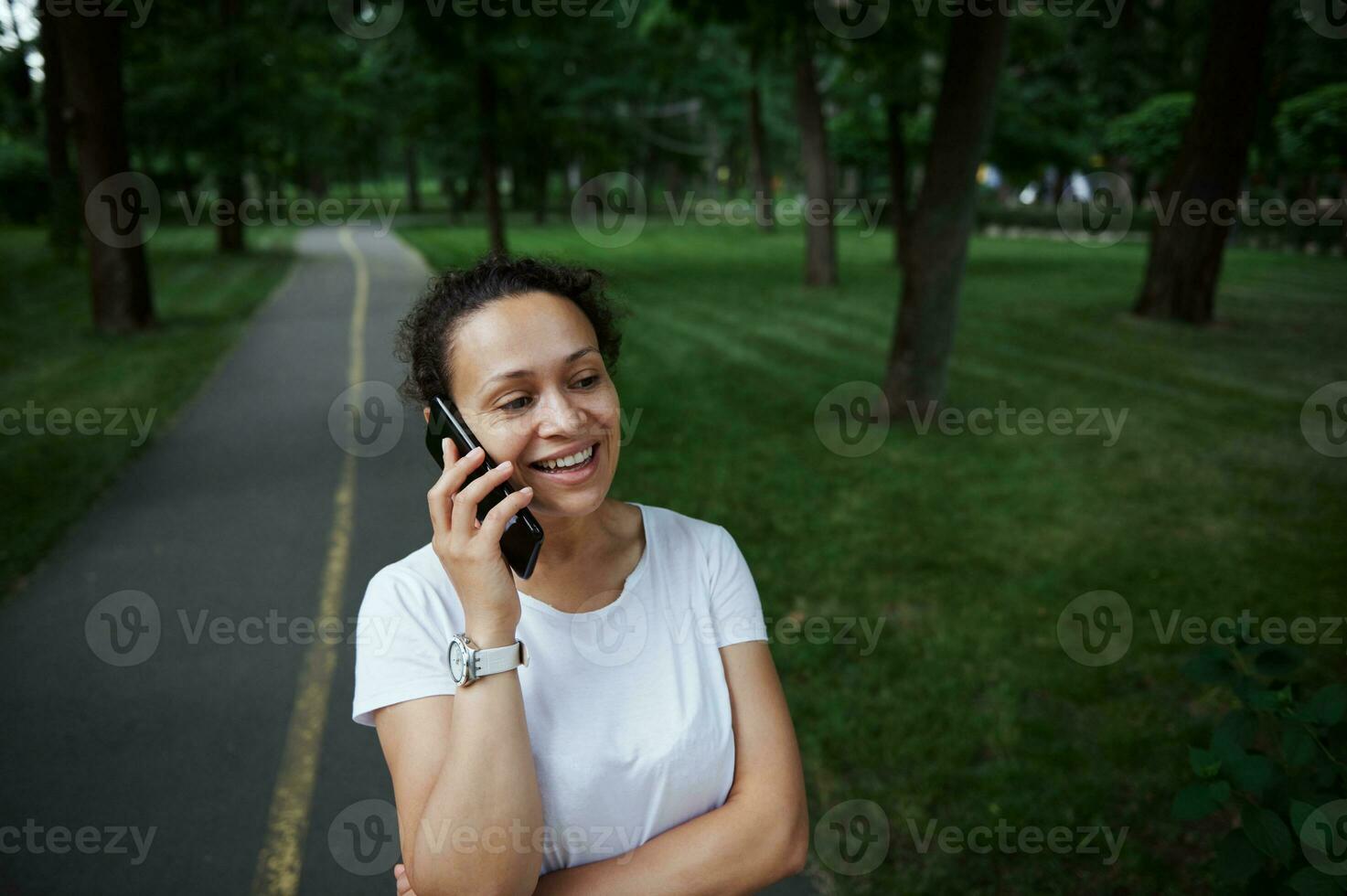 Beautiful female tourist in white t-shirt, talks on mobile phone while strolling the alley of a city park o summer day photo