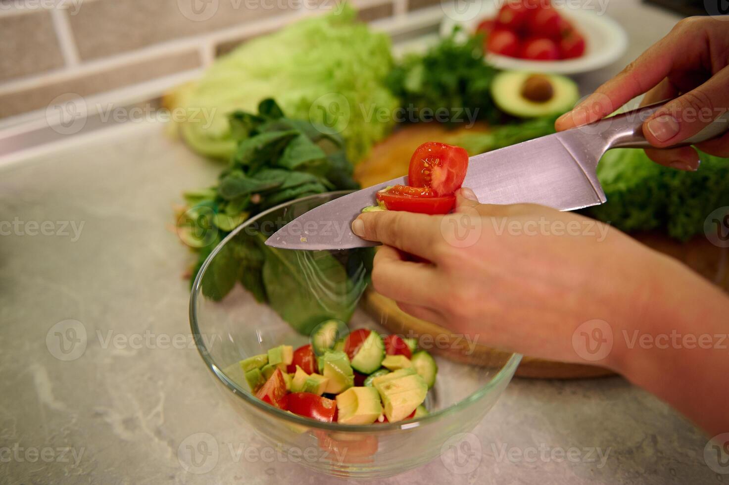 High angle view of female hands cutting vegetables with kitchen knife and putting them on glass transparent bowl. Vegan and healthy lifestyle concepts. Preparing raw vegan salad photo