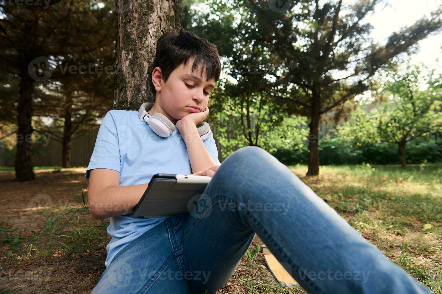 hermoso Hispano adolescente colegio chico haciendo deberes en digital tableta, sentado relajado en el parque. niños. en línea educación foto